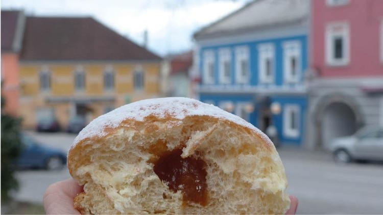 Im Vordergrund wird ein halbierter Krapfen gehalten, im Hintergrund stehen bunde Häuser.