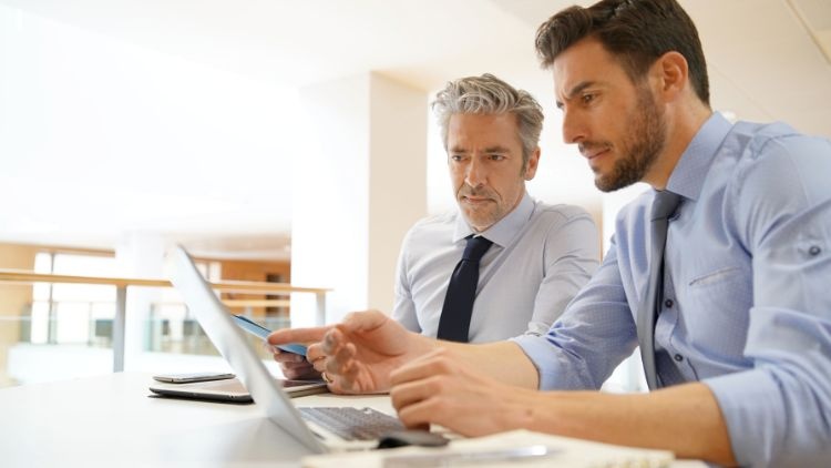 © goodluz | stock.adobe.com Zwei Personen in Hemden und mit Krawatten sitzen nebeneinander an einem Tisch. Vor ihnen steht ein aufgeklappter Laptop, auf den beide klicken. Neben dem Laptop sind ein Notizbuch und ein Tablet.